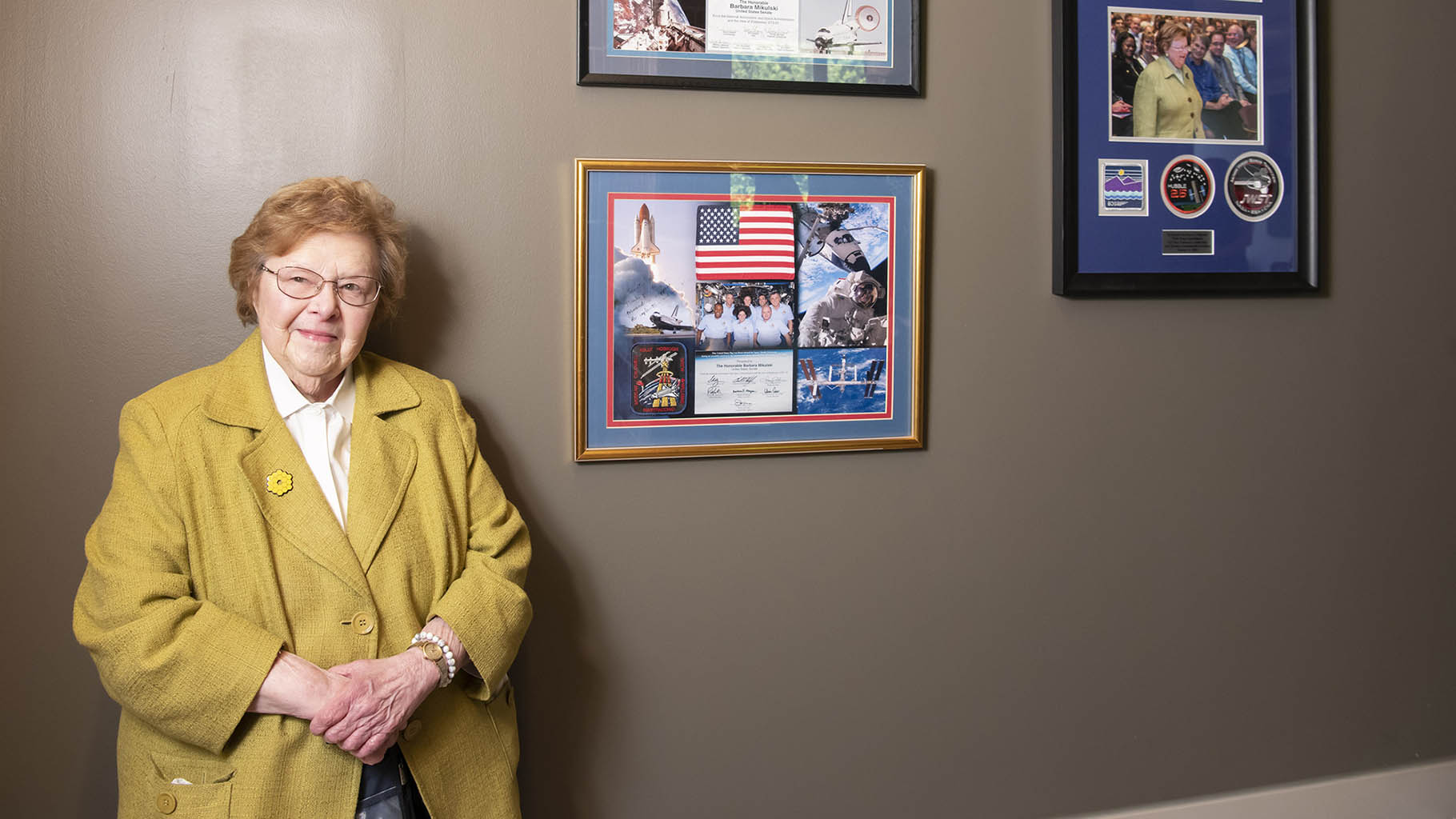 Photograph of Senator Barbara A. Mikulski in a yellow jacket with a white shirt taken July 12, 2022, when she visited the Space Telescope Science Institute to celebrate the public release of the first images from the James Webb Space Telescope. Mikulski is standing in front of framed memorabilia from her donated collection that now hangs in the Muller building on the Johns Hopkins Homewood campus. 