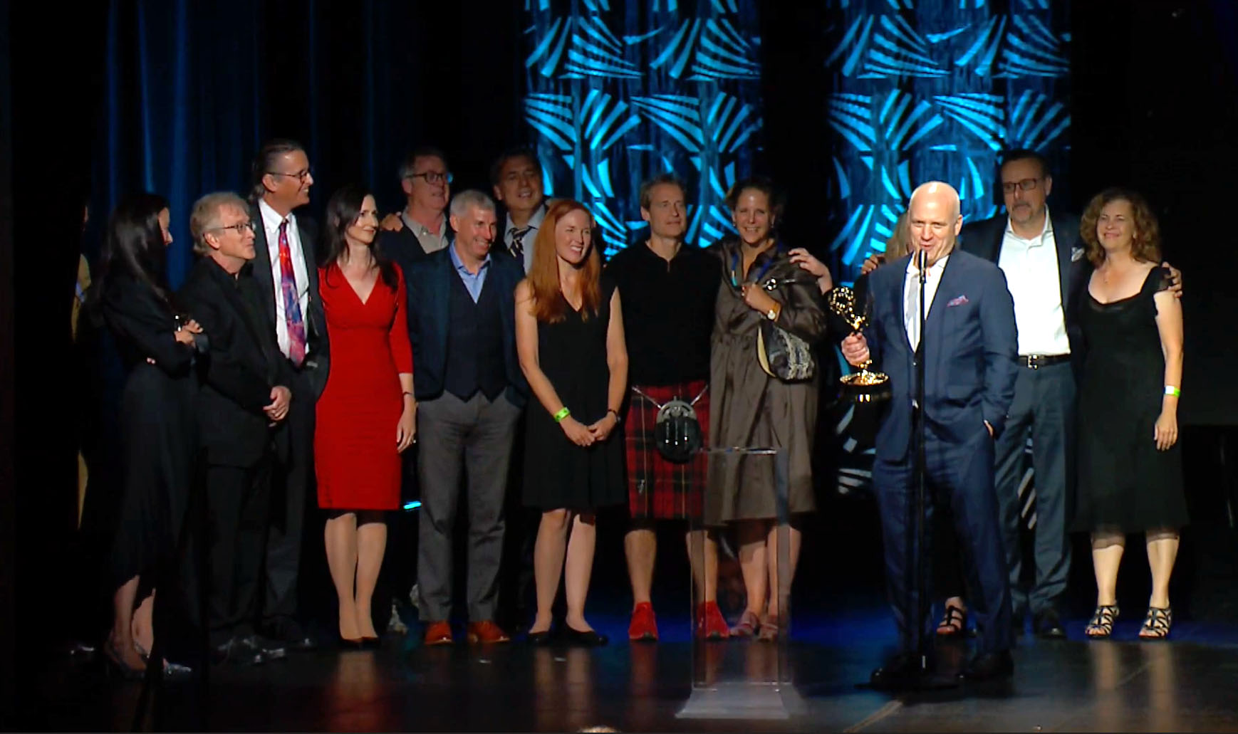 Nathaniel Kahn stands in front of a mic with the Emmy in his hand accepting the award. Behind him are about 15 people who worked on or were portrayed in the film "the Hunt for Planet B."