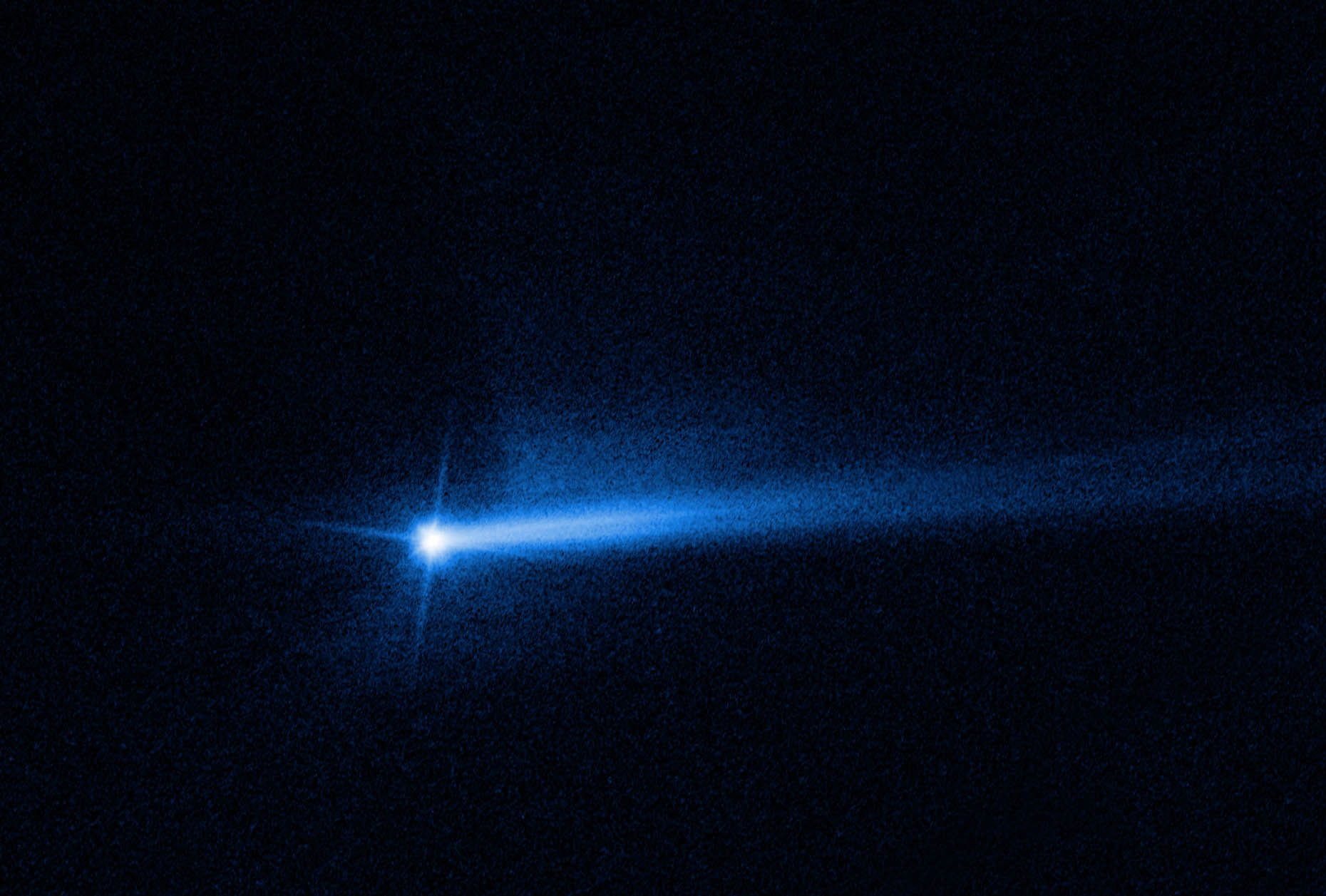 A bright blue spot is at the left-center of the image, which has a black background. The spot is the Didymos-Dimorphos system after impact from the DART spacecraft. The center bright spot has 3 diffraction spikes extending from its core at the 1 o’clock, 7 o’clock, and 10 o’clock positions. There is a small amount of dusty haze just below the southern pole of the center dot. Two tails of ejecta that appear as white streams of material extend out from the center at the 2 o’clock and 3 o’clock positions.