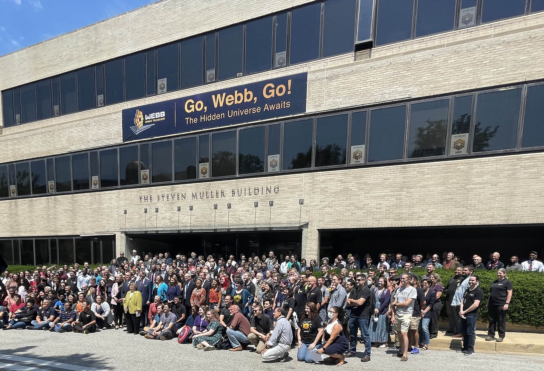 A crowd of people outside of the STSc. building cheer the first science results of JWST. A banner with the text "Go, Webb, Go!" hangs on the building above them.