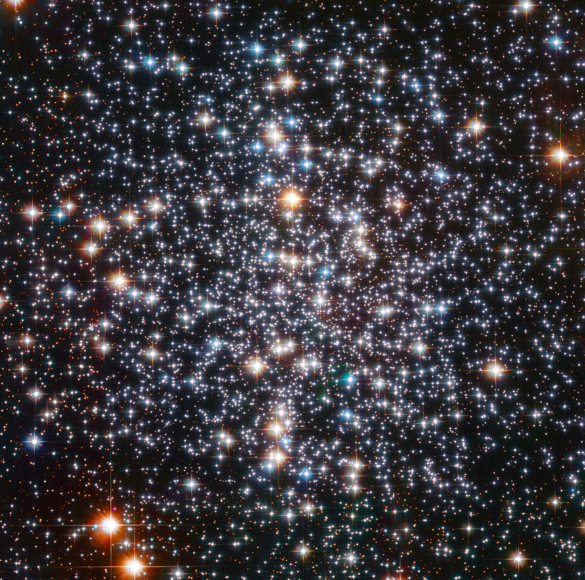 Thousands of bright points of light on a black background fill the field of view. They appear somewhat more concentrated near the center of the image. They have a variety of colors, with the most prominent stars appearing blue or yellow-orange. The brighter stars also display four diffraction spikes.