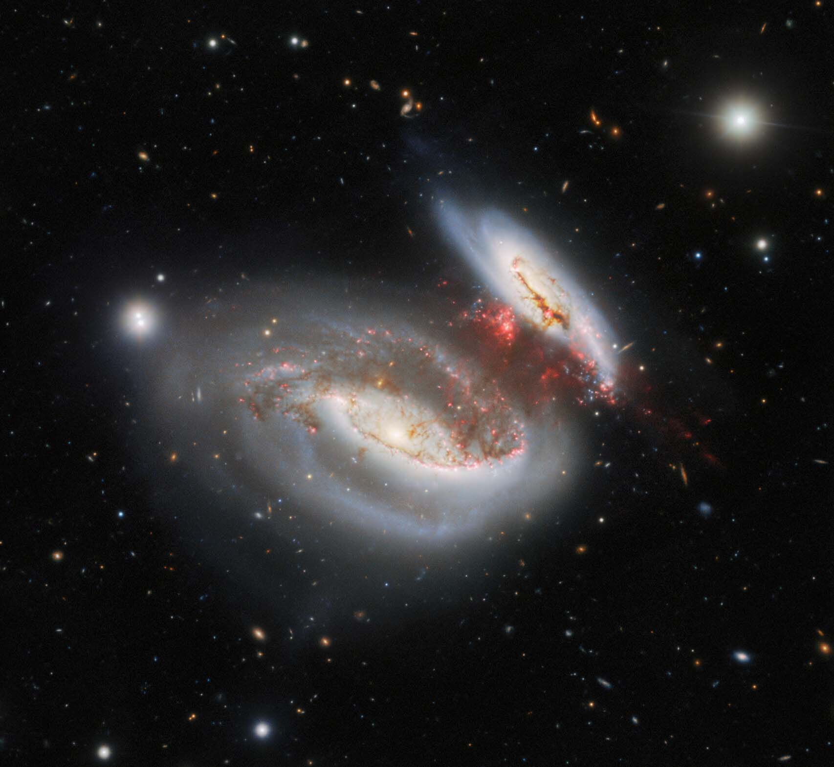 2 bright pinwheel galaxies move close together with bright red spots between them on a black background speckled with distant galaxies.