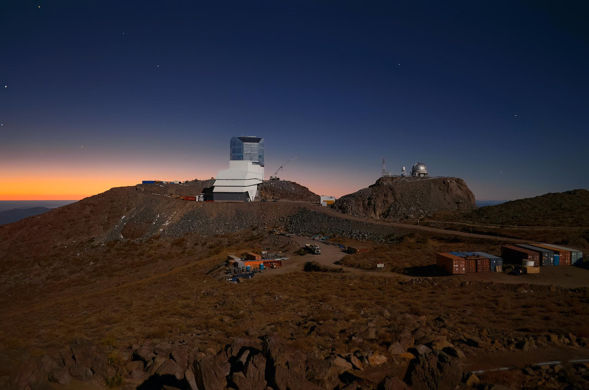 A white observatory building with a shiny silver dome sits on a rocky site under a dark late-twilight sky. A second small dome sits on a neighboring hill. The sky fills the top half of the image and is a uniform dark blue, with a hint of orange on the left horizon. Tiny pinpricks of stars are scattered in the sky. The brown, rocky desert landscape fills the bottom half of the image, darkened so that details are hard to pick out. A collection of shipping containers are lined up side by side in the lower right.