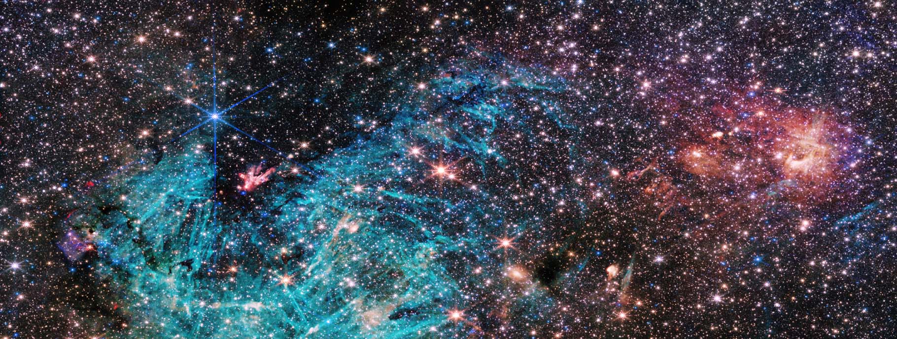 a funnel-shaped region of space appears darker than its surroundings with fewer stars. It is wider at the top edge of the image, narrowing towards the bottom. Toward the narrow end of this dark region a small clump of red and white appears to shoot out streamers upward and left. A large, bright cyan-colored area surrounds the lower portion of the funnel-shaped dark area, forming a rough U shape. The cyan-colored area has needle-like, linear structures and becomes more diffuse in the center of the image. The right side of the image is dominated by clouds of orange and red, with a purple haze.