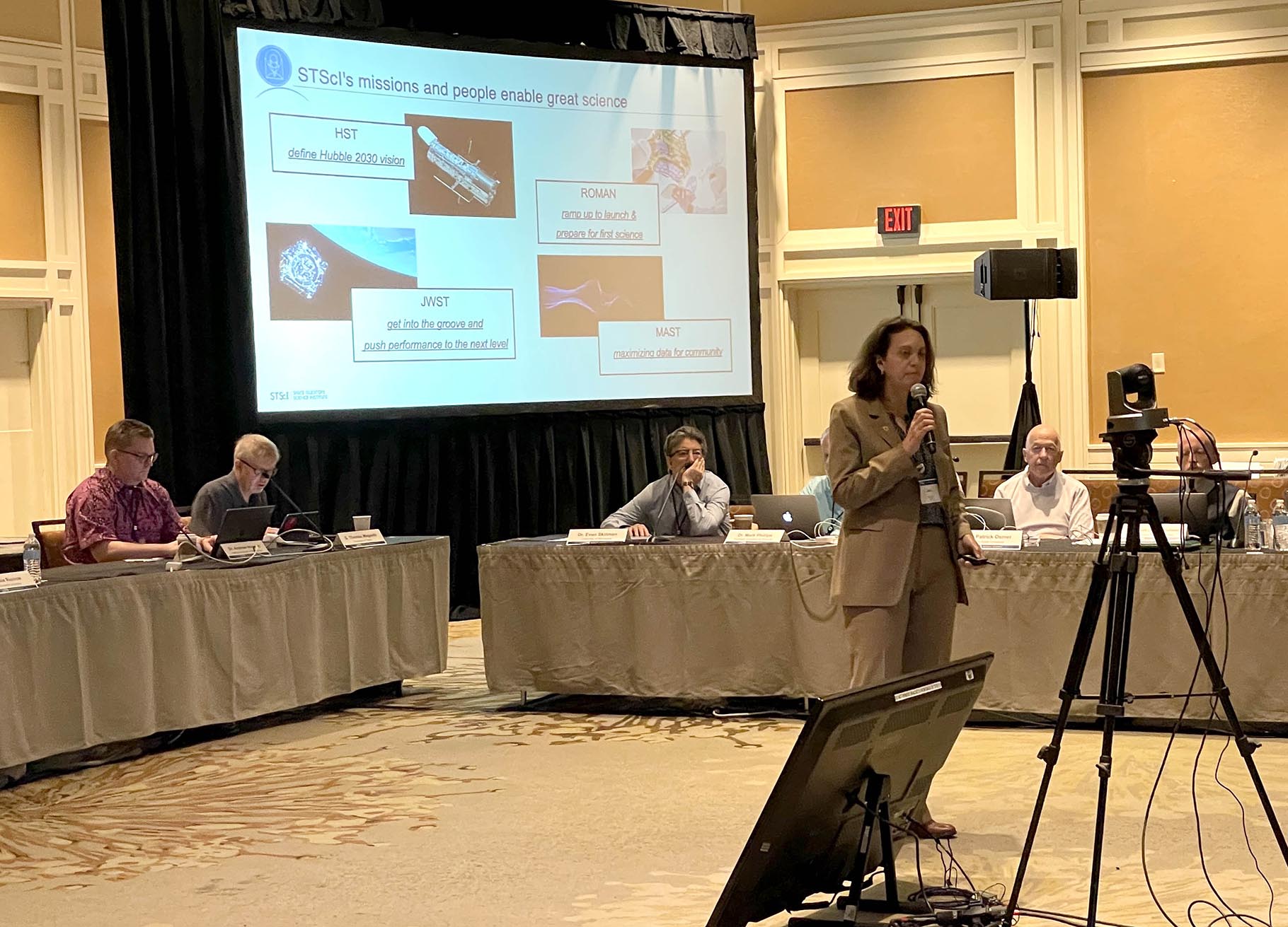 Jen Lotz with mic in hand talking in front of a screen showing space telescopes.