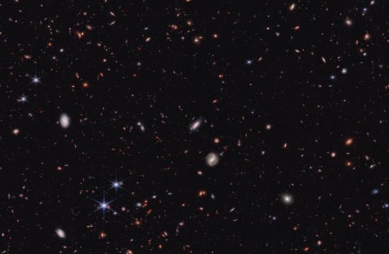Hundreds of small galaxies against the black background of space. Several white spiral galaxies are near image center. Most of the galaxies are various shades of orange and red, and many are too tiny to discern a shape. A handful of foreground stars show Webb's six diffraction spikes.