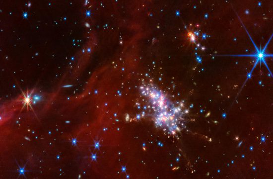 At center right is a compact star cluster composed of luminous red, blue, and white points of light. Faint jets with clumpy, diffuse material extend in various directions from the bright cluster. Above and to the right is a smaller cluster of stars. Translucent red wisps of material stretch across the scene, though there are patches and a noticeable gap in the top left corner that reveal the black background of space. Background galaxies are scattered across this swath of space, appearing as small blue-white and orange-white dots or fuzzy, thin disks. There are two noticeably larger points, foreground stars, with diffraction spikes: an orange-white point on the left, and a blue-white point in the top right.