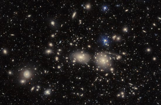 A multitude of stars and galaxies on a dark background. In the center are two brighter galaxies with a yellow glow around them.
