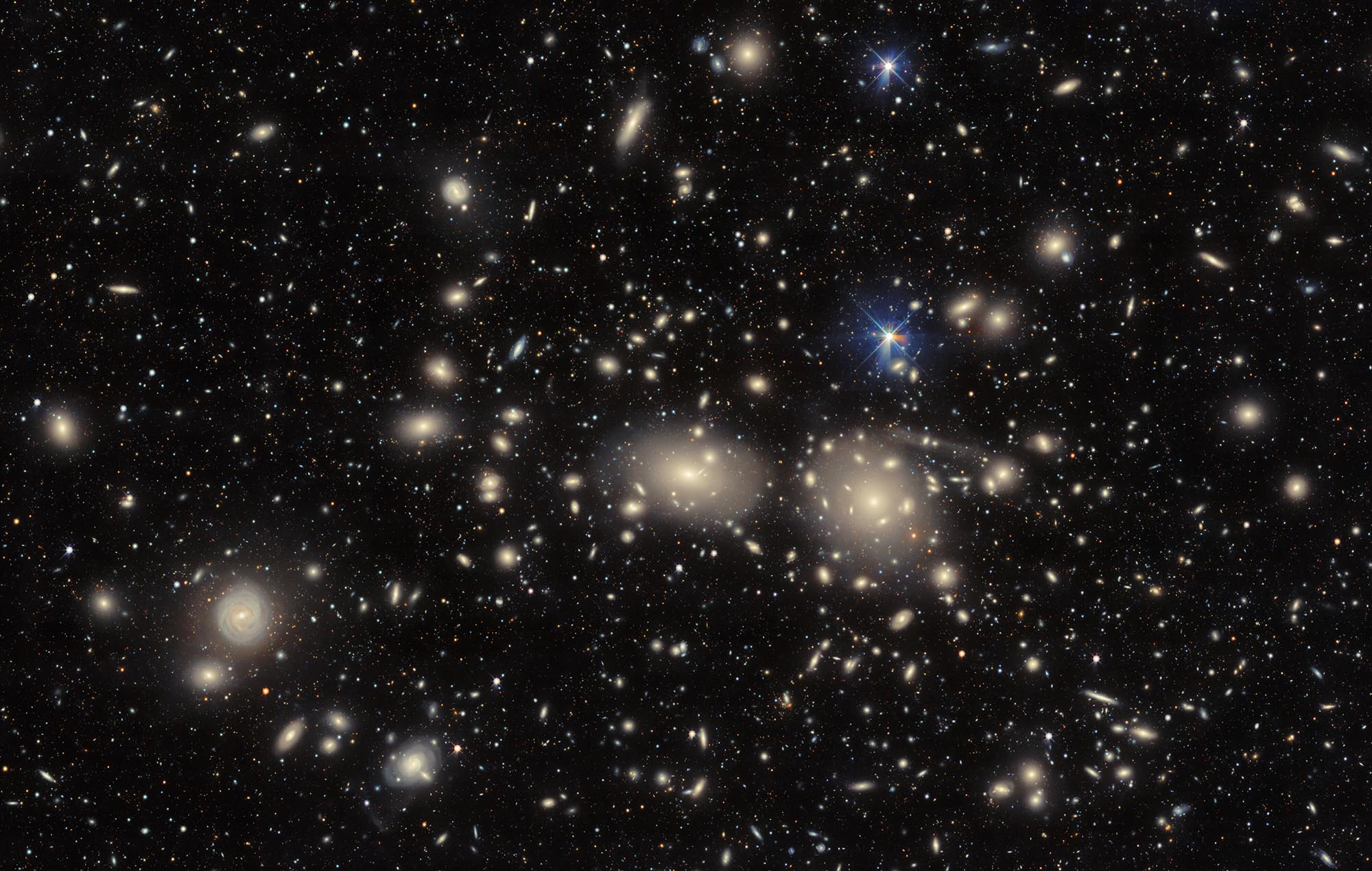 A multitude of stars and galaxies on a dark background. In the center are two brighter galaxies with a yellow glow around them.
