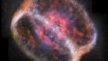 What looks like a single large, bright star but is two shines with bright purple diffraction spikes at the center of a large, diffuse cylinder of gas and dust that is tipped to the right. At the center is a bright pink clumpy cloud that takes up about 25% of the view. The pink region has some holes. Beyond that are two large rings tipped at a 60-degree angle that mark the ends of the cylinder. They appear joined at top left and bottom right. The edges form shallow V-shapes that go inward. The rings appear orange at top left and bottom right, and are blue at bottom and center right. There is diffuse orange material around the body. The black background of space is speckled with tiny stars and galaxies mostly in blues and yellows. A bigger blue star with spikes is just below and to the left of the central stars, but it is slightly smaller. Areas Webb did not observe appear black along the top edges, a thin vertical near the nebula at top left, and at the bottom left and right corners.