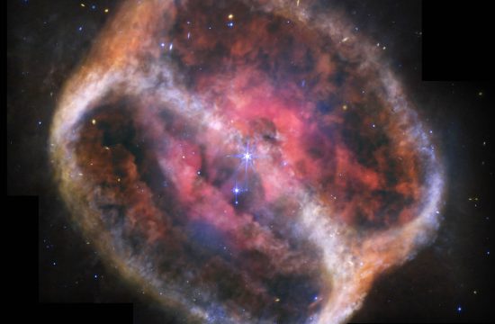 What looks like a single large, bright star but is two shines with bright purple diffraction spikes at the center of a large, diffuse cylinder of gas and dust that is tipped to the right. At the center is a bright pink clumpy cloud that takes up about 25% of the view. The pink region has some holes. Beyond that are two large rings tipped at a 60-degree angle that mark the ends of the cylinder. They appear joined at top left and bottom right. The edges form shallow V-shapes that go inward. The rings appear orange at top left and bottom right, and are blue at bottom and center right. There is diffuse orange material around the body. The black background of space is speckled with tiny stars and galaxies mostly in blues and yellows. A bigger blue star with spikes is just below and to the left of the central stars, but it is slightly smaller. Areas Webb did not observe appear black along the top edges, a thin vertical near the nebula at top left, and at the bottom left and right corners.