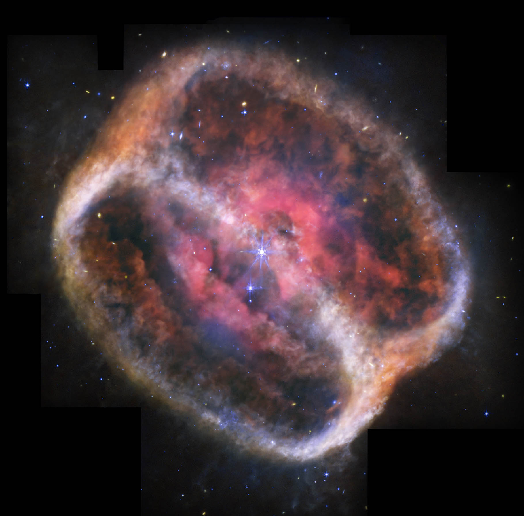 What looks like a single large, bright star but is two shines with bright purple diffraction spikes at the center of a large, diffuse cylinder of gas and dust that is tipped to the right. At the center is a bright pink clumpy cloud that takes up about 25% of the view. The pink region has some holes. Beyond that are two large rings tipped at a 60-degree angle that mark the ends of the cylinder. They appear joined at top left and bottom right. The edges form shallow V-shapes that go inward. The rings appear orange at top left and bottom right, and are blue at bottom and center right. There is diffuse orange material around the body. The black background of space is speckled with tiny stars and galaxies mostly in blues and yellows. A bigger blue star with spikes is just below and to the left of the central stars, but it is slightly smaller. Areas Webb did not observe appear black along the top edges, a thin vertical near the nebula at top left, and at the bottom left and right corners.