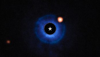 An image of a nearby star and its vicinity. The star itself has been blocked out and its bright light has been removed, appearing black. A dashed circle with a star symbol at the center of the image marks the star’s location. A fuzzy blue disk surrounds the star. An orange spot, near the star and inside this disk around 2 o’clock, is identified as a planet orbiting the star. A fainter orange spot at the lower left edge marks a distant star.