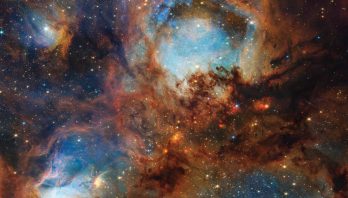 A section of the Cat’s Paw Nebula, a local star-forming region composed of gas, dust, and young stars. Four roughly circular areas are toward the center of the frame: a small oval toward the top left, a large circle in the top center, and two ovals at bottom left and right. Each circular area has a luminous blue glow, with the top center and bottom left areas the brightest. Brown-orange filaments of dust, which vary in density, surround these four bluish patches and stretch toward the frame’s edges. Small zones, such as to the left and right of the top-center blue circular area, appear darker and seemingly vacant of stars. Toward the center are small, fiery red clumps scattered among the brown dust. Many small, yellow-white stars are spread across the scene, some with eight-pointed diffraction spikes that are characteristic of Webb. A few larger blue-white stars with diffraction spikes are scattered throughout, mostly toward the top left and bottom right. In the top right corner is a bright red-orange oval.