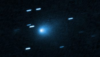 At the center of the image is a comet that appears as a teardrop-shaped bluish cocoon of dust coming off the comet’s solid, icy nucleus and seen against a black background. The comet appears to be heading to the bottom left corner of the image. About a dozen short, light blue diagonal streaks are seen scattered across the image, which are from background stars that appeared to move during the exposure because the telescope was tracking the moving comet.