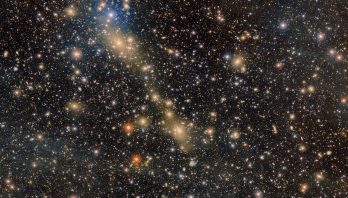 A glittering diagonal group of glowing yellow galaxies shine out of the image filled with glowing points of light.