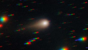 A glowing ball with a gold halo and tail streaks to the right in the center of this image. The black background is cover with rainbow colored streaks throughout the image.