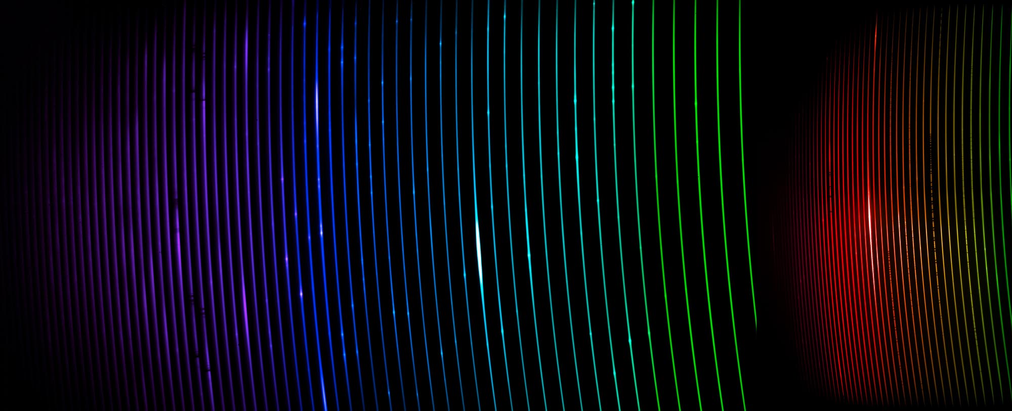 Slightly curved thin luminous lines from purple to turquoise to green to red and then yellow, run vertically over on a black background.