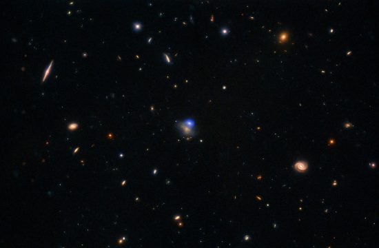 Bright reds, yellows and orange points of lights are scattered on a black background Some are obviously galaxies with small pinwheel shape and others are just points of light. In the Center is a bright blue glowing object next to a fainter orange one.