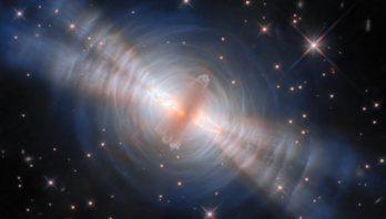 In the image center, an opaque oval cloud of gray gas aligned from 1 o’clock to 7 o’clock hides a star. Two strong beams of light from the star emerge from large holes in both sides of the cloud, forming narrow cones extending toward 10 o’clock and 4 o’clock. The central cloud is surrounded by concentric, wispy shells of gas illuminated by the star’s light. The shells reflect extra light where they’re hit by the twin beams. A crowd of smaller stars with cross-shaped spikes over them surrounds the nebula on a black background.