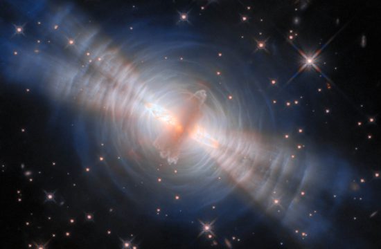 In the image center, an opaque oval cloud of gray gas aligned from 1 o’clock to 7 o’clock hides a star. Two strong beams of light from the star emerge from large holes in both sides of the cloud, forming narrow cones extending toward 10 o’clock and 4 o’clock. The central cloud is surrounded by concentric, wispy shells of gas illuminated by the star’s light. The shells reflect extra light where they’re hit by the twin beams. A crowd of smaller stars with cross-shaped spikes over them surrounds the nebula on a black background.