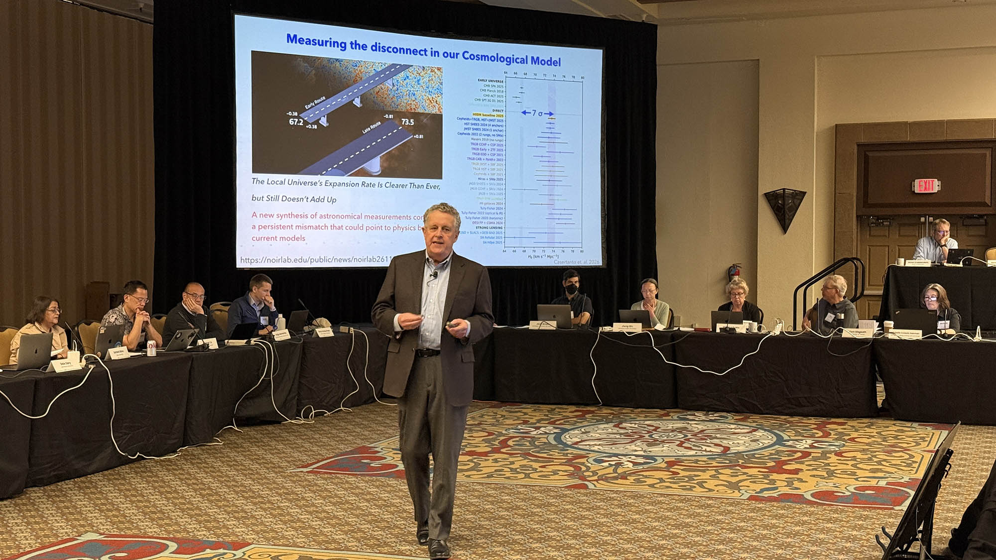 Man in a suit walks towards the camera while speaking in a large conference room with a large screen showing a science slide. Attendees watch from behind him.
