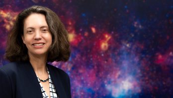 A photograph of a woman with shoulder-length dark hair wearing a dark blazer, checkered shirt, and necklace against a colorful background image of a nebula.