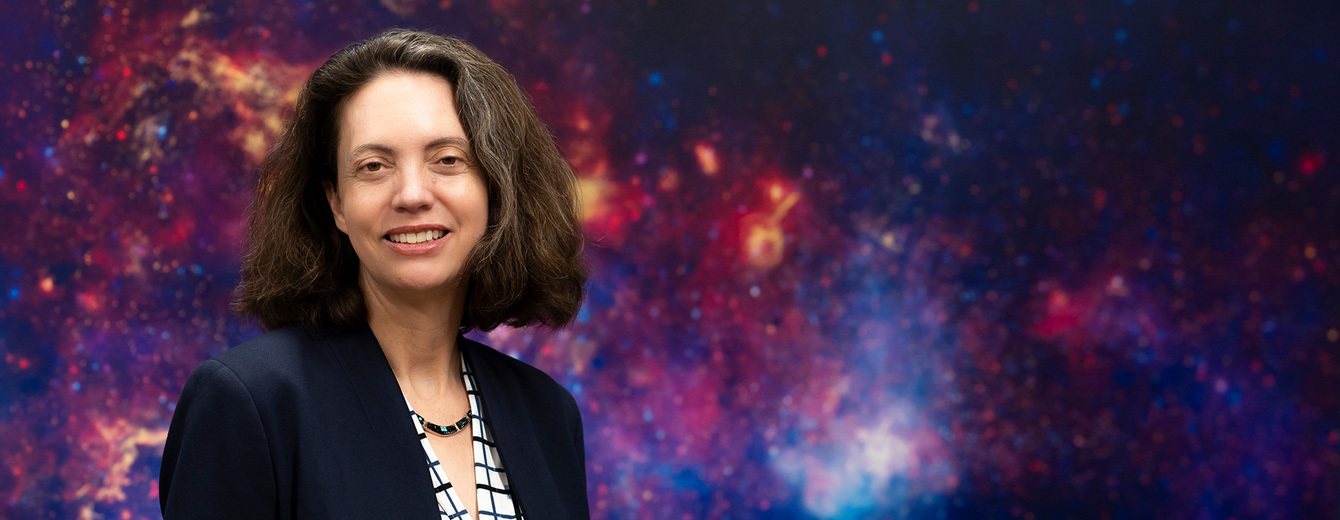 A photograph of a woman with shoulder-length dark hair wearing a dark blazer, checkered shirt, and necklace against a colorful background image of a nebula.