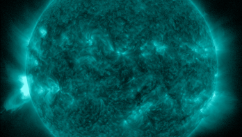 A full-disk extreme ultraviolet visualization of the Sun rendered in a brilliant, textured cyan-teal hue against a black background. The solar surface is covered in intricate, mottle patterns with darker filimentary structures and multiple smaller bright active regions. On the lower-left edge, a blindingly bright white-cyan eruption—a long-duration M-class solar flare—is actively exploding from the limb, with brilliant hot plasma and detailed magnetic loops extending into the void of space, originating from a region just beyond the Sun's edge.